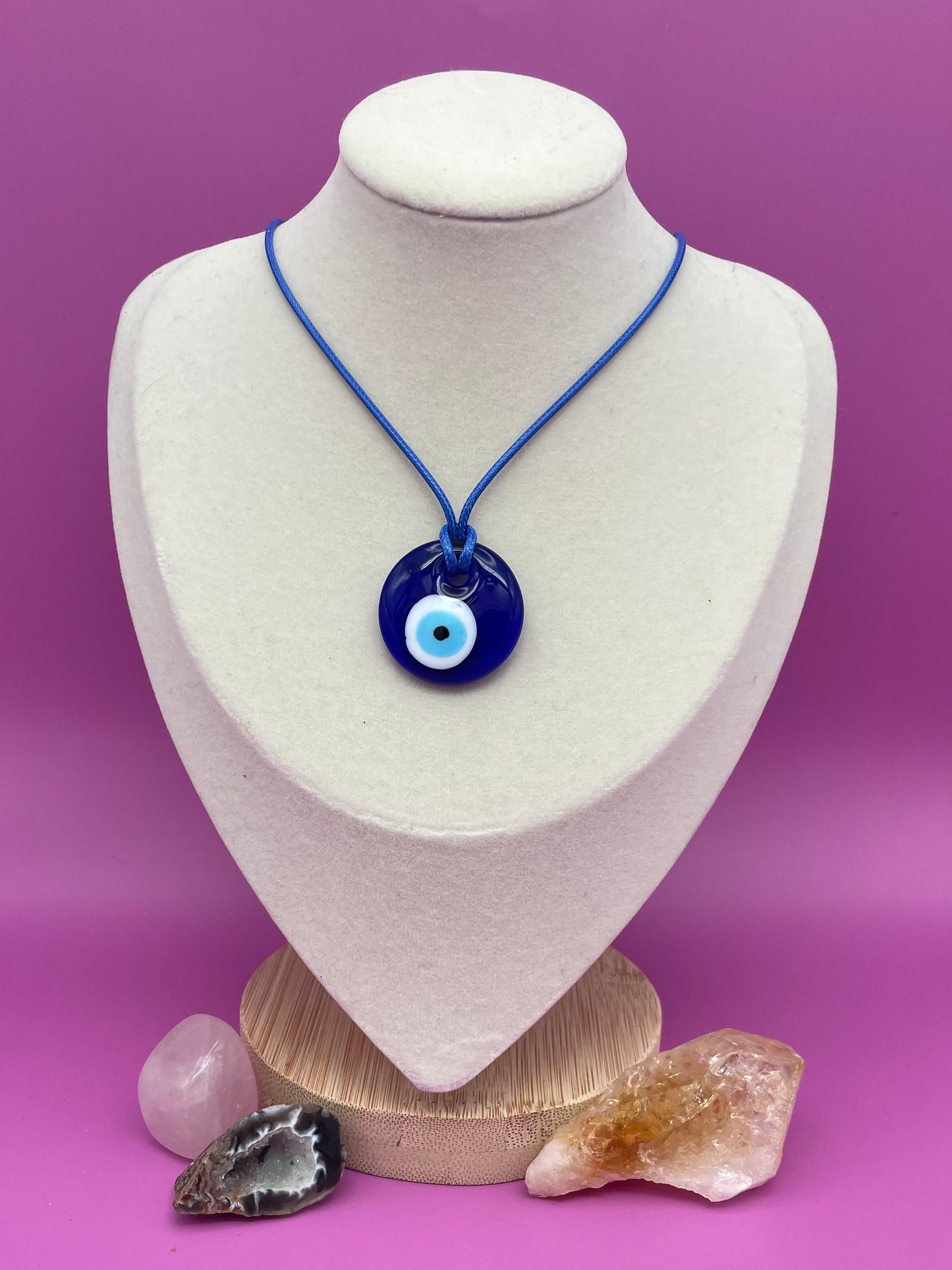 Blue glass evil eye pendant necklace with blue cord on a white mannequin bust against a purple background